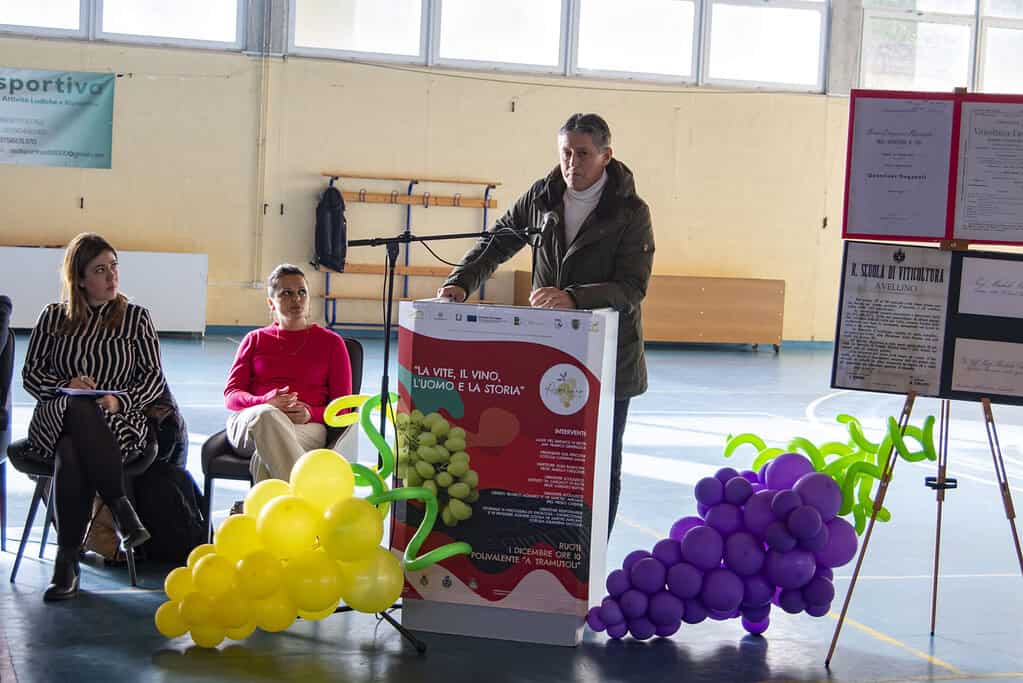Degustazione di vino in palestra con pubblico e presentatore, simbolo della connessione tra vite, vi.